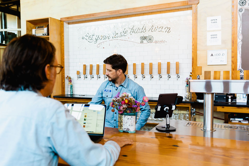person serves beer behind a bar, with another person sitting at the bar on their computer