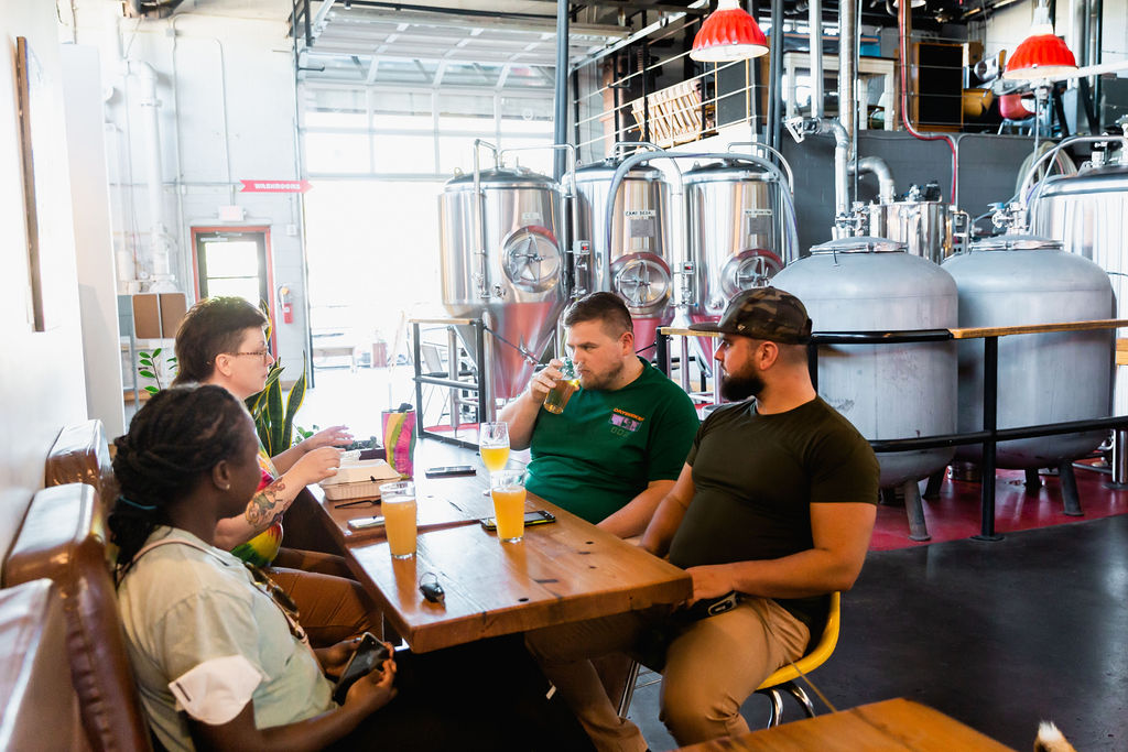 A group of 4 people sitting at a table, one drinking a beer, with beer fermenting tanks sitting behind them.