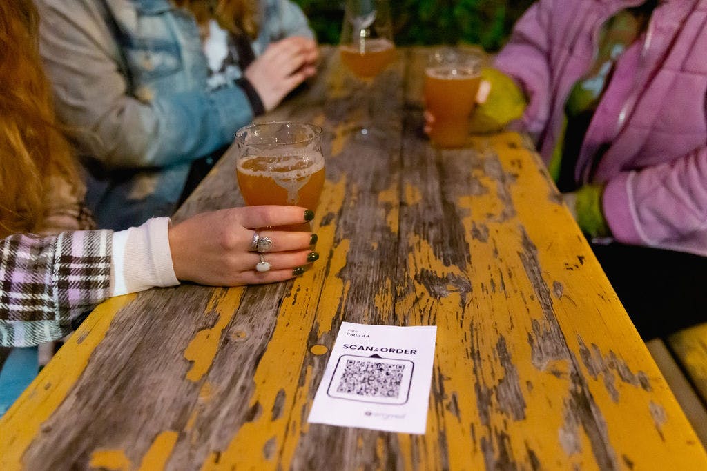 Hands hold cups of beer on a wooden table with a QR code for ordering