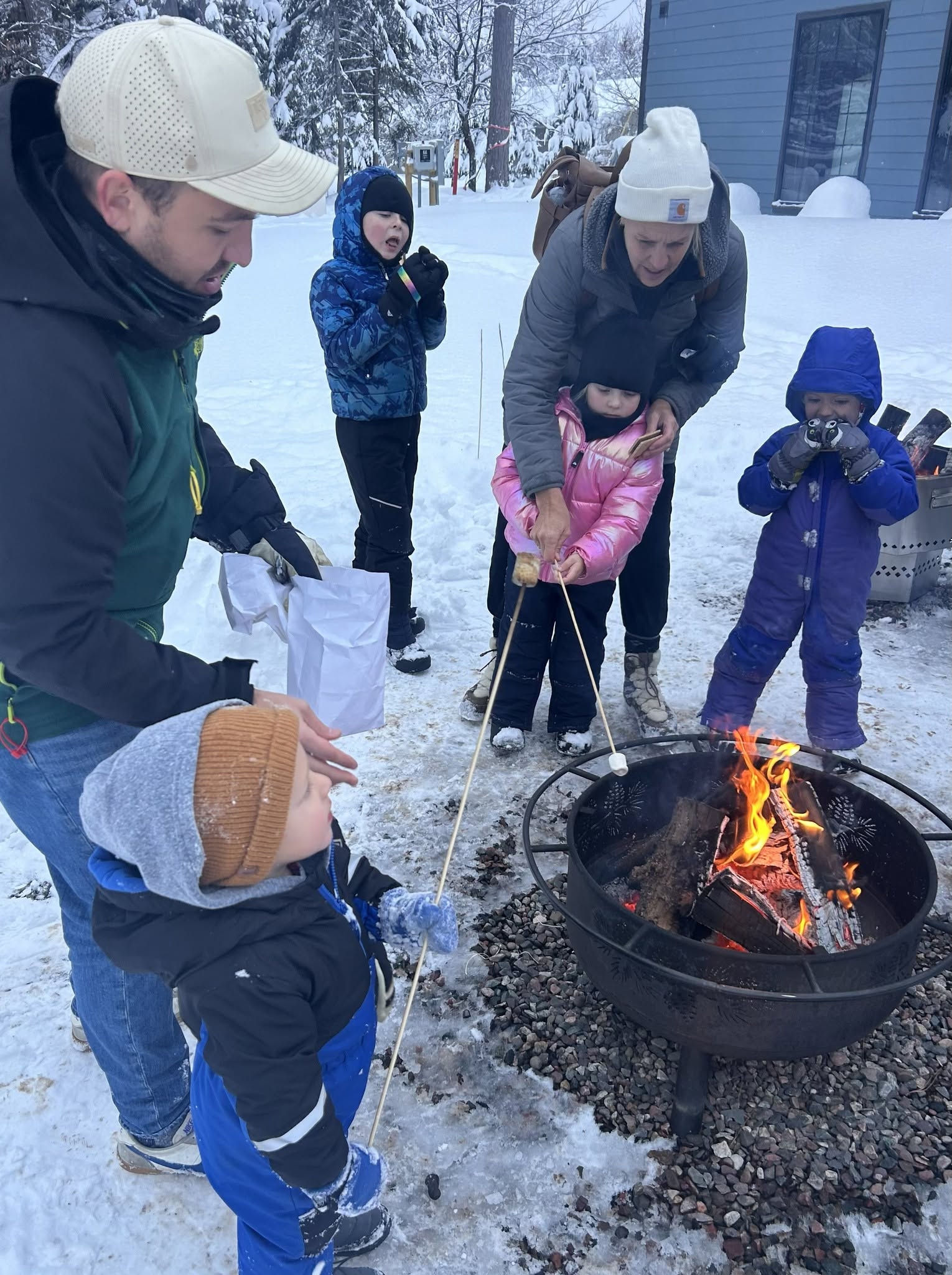 snowy firepit with a dad helping 2 children roast marshmallows