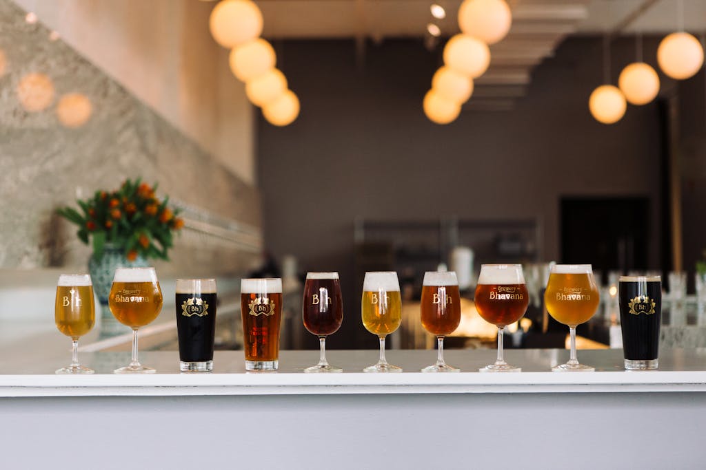 A row of various beer glasses on a bar, showcasing different types and colors of beer, with a floral arrangement in the background.