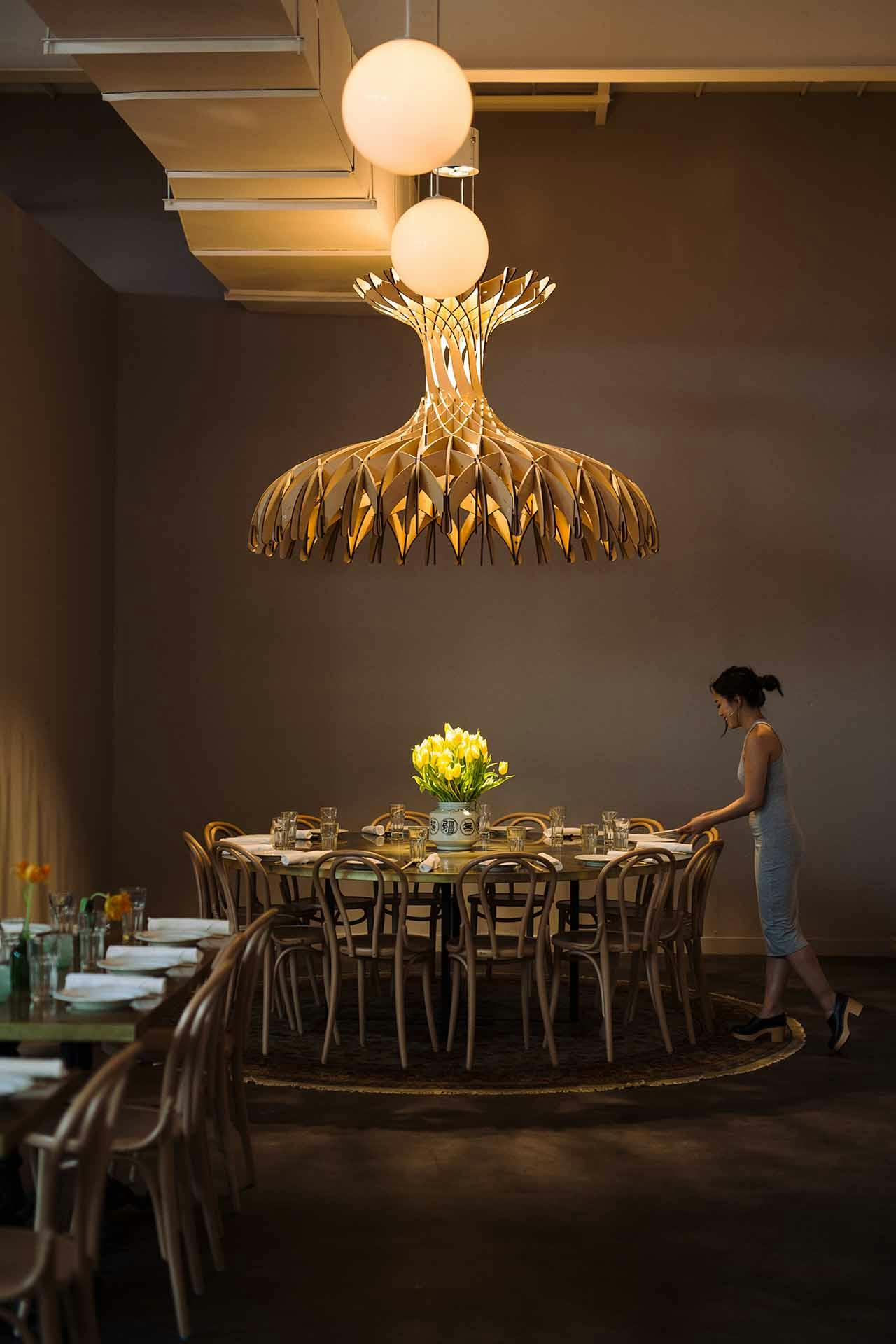 a woman sets a round table under an intricate wooden light fixture
