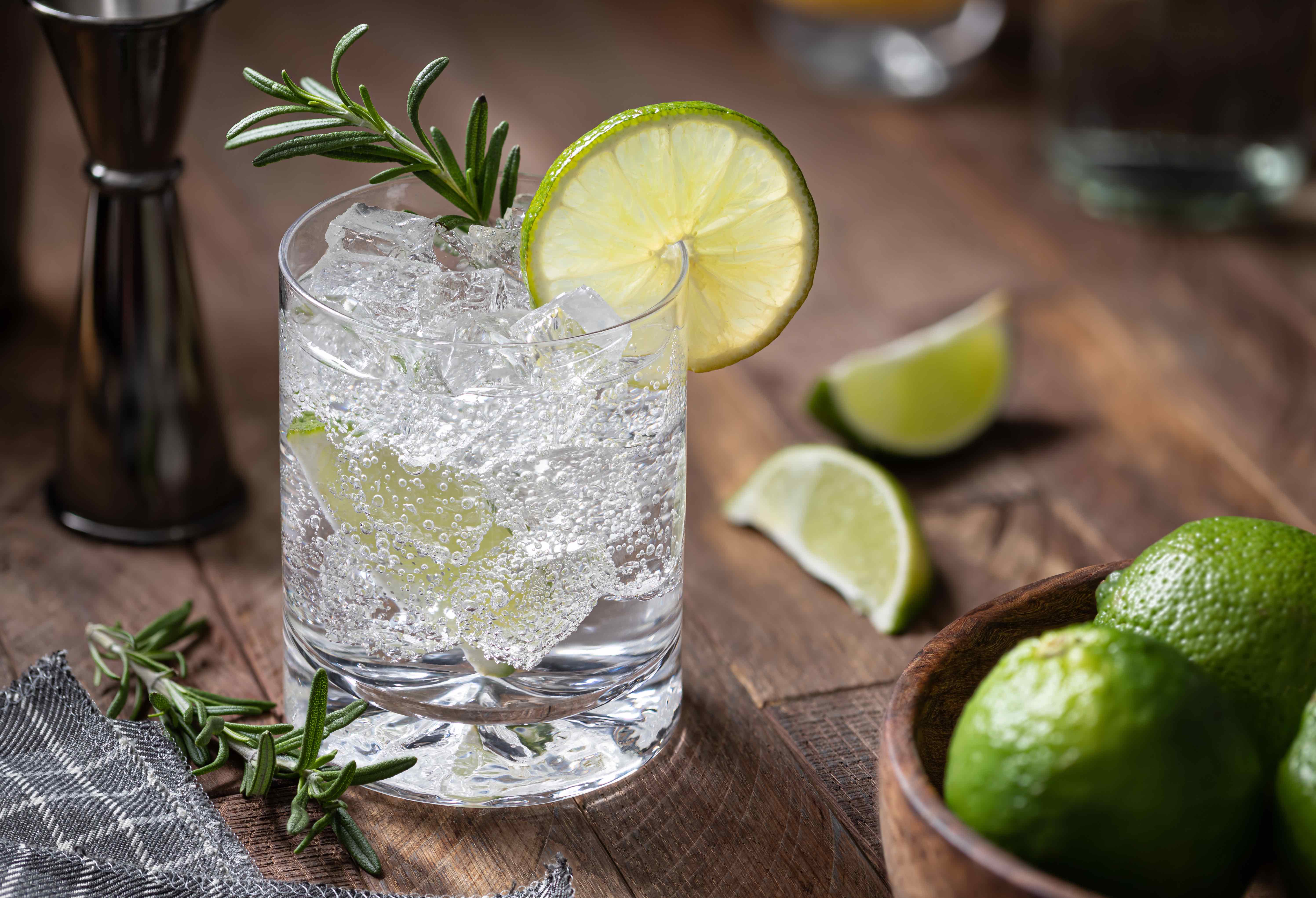 Gin and tonic cocktail with lime slices, rosemary and ice on a rustic wooden table