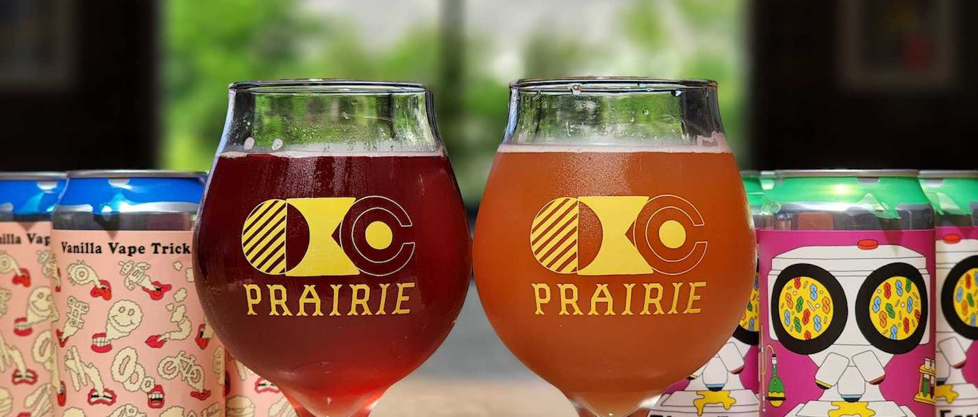 Two glasses of Prairie beers in logo glasses on a wooden table, with 6-packs of cans behind