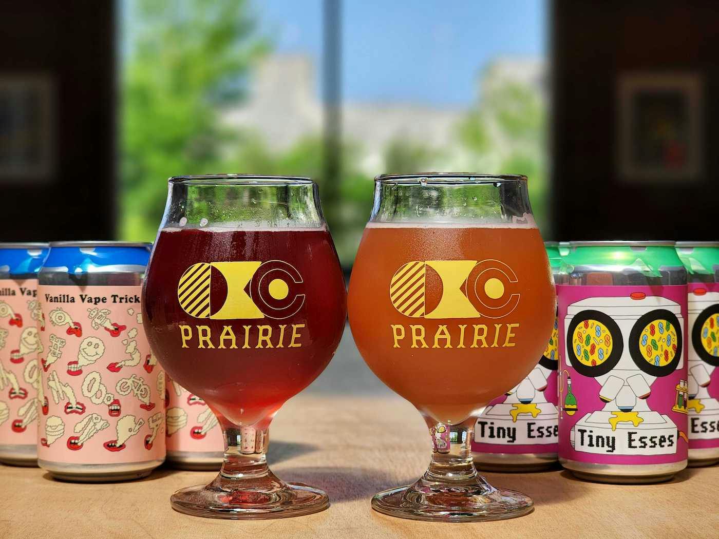 Two glasses of Prairie beers in logo glasses on a wooden table, with 6-packs of cans behind