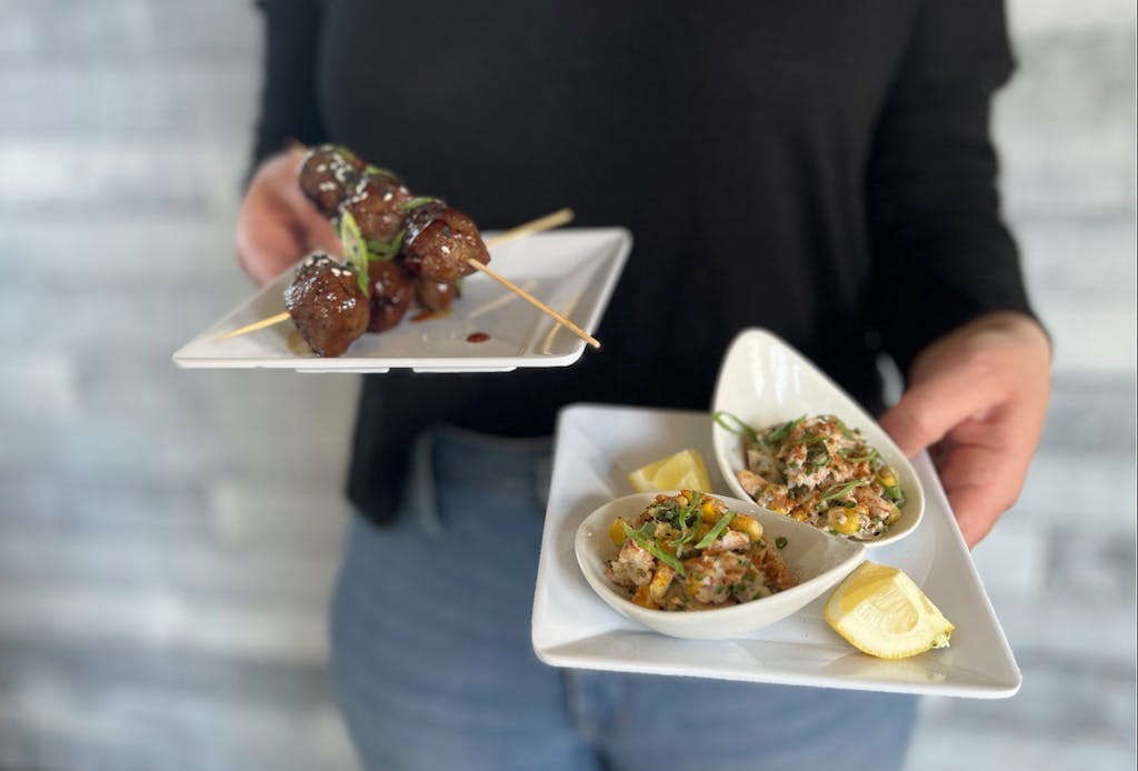 The Galley at Rising Tide Appetizers Person holding two square white plates. Foreground plate hold two small bowls of crab salad and lemon wedges, the second plate holds two skewers of meatballs.