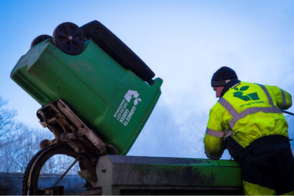 Photograph of Garbage to Garden picking up a commercial bin.  Green bin is lifted in the air and emptying into a turck out of frame.  There is a man in a neon yellow jacket with Garbage to Garden logo leaving over the truck. 