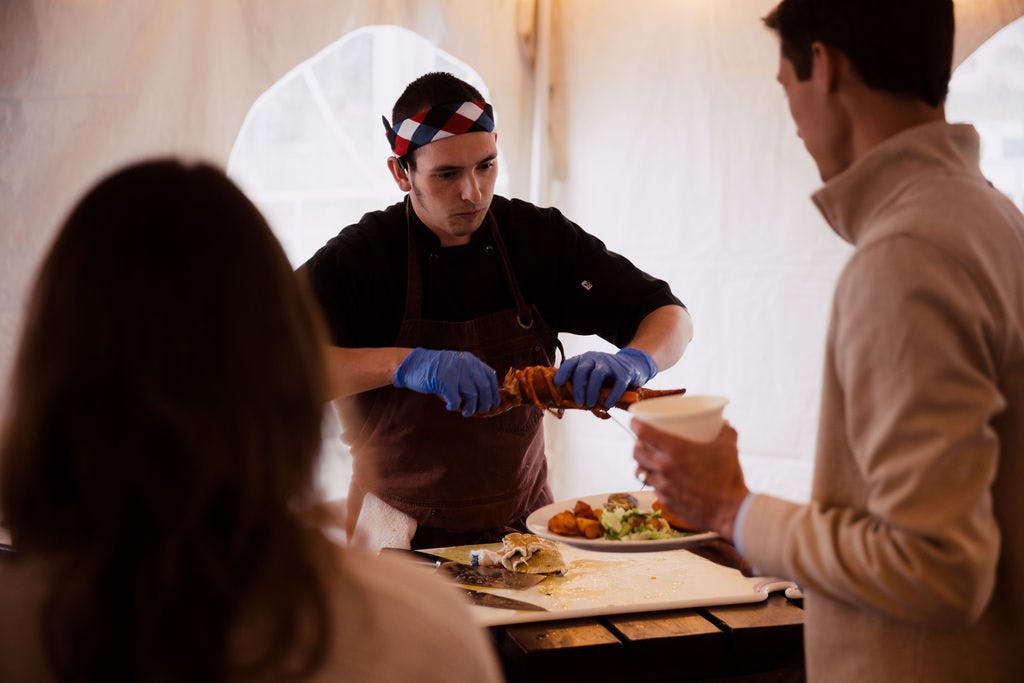 H-S_Rehearsal(76of110) Rising Tide cook Kyle serving freshly baked lobster to a man in a white shirt under the tent during an event.