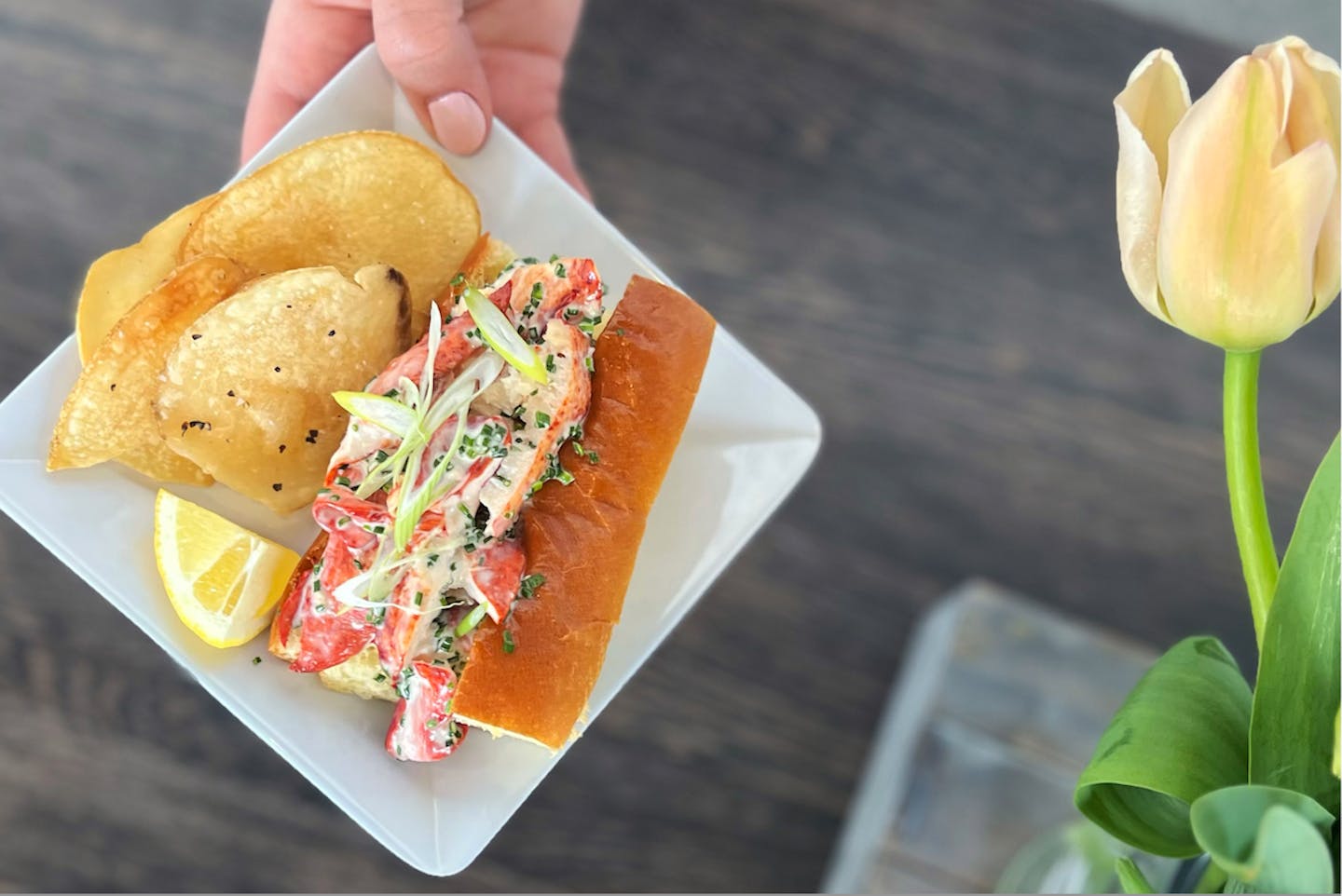 Hand holding a white plate with a lobster roll, chips, and lemon wedge. There is a soft yellow tulip on the far right side of the image.