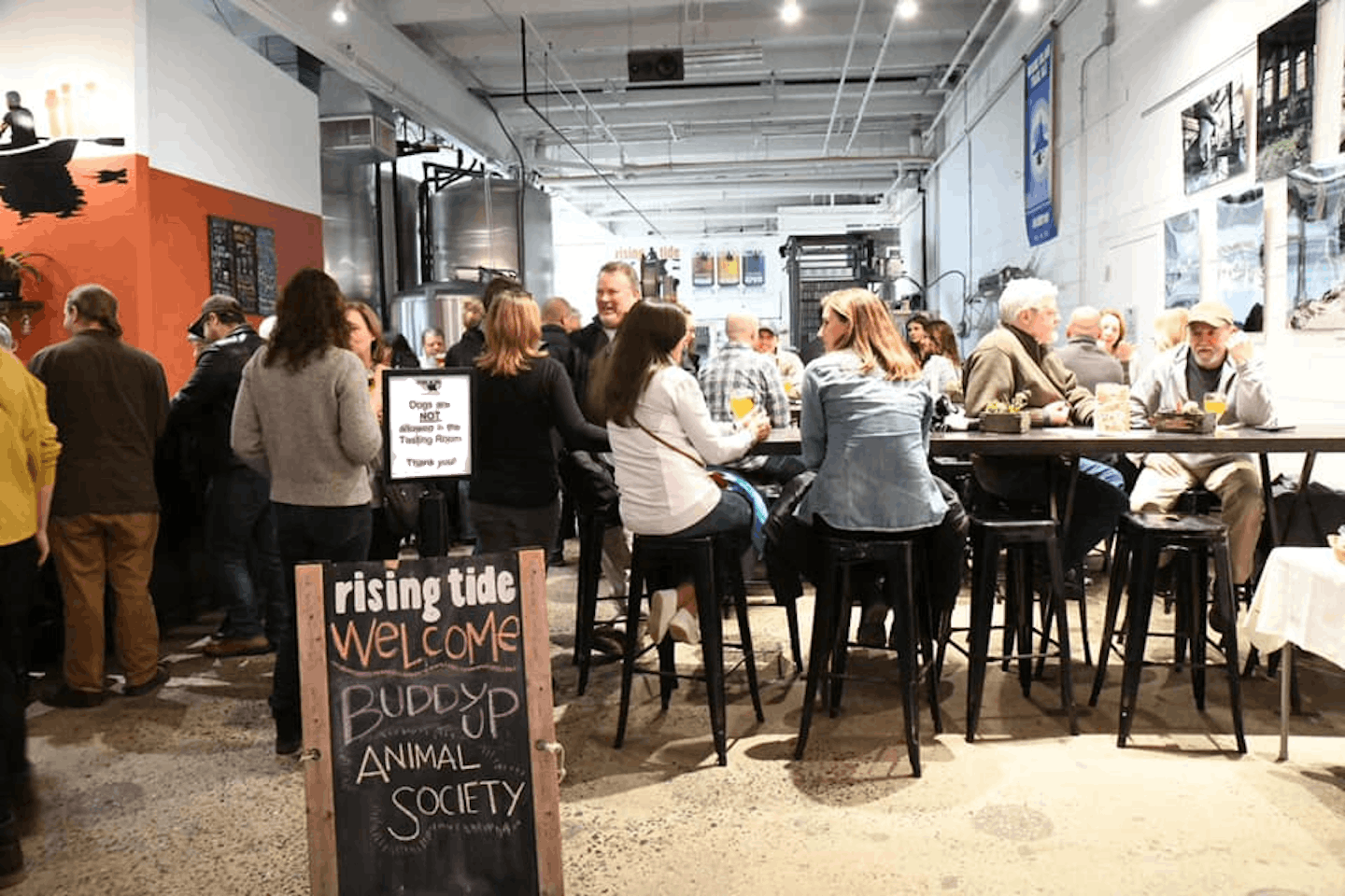 Photograph of the interior of the tasting room.  There is a chalkbaord sign that ready Buddy Up Animal Society in the foreground and people mingling at the bar and tables in the background