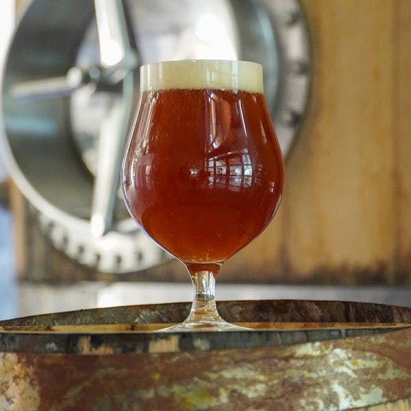 A red beer in a clear, round glass on top of a wooden barrel. Blurred in the background is a larger barrel for brewing.