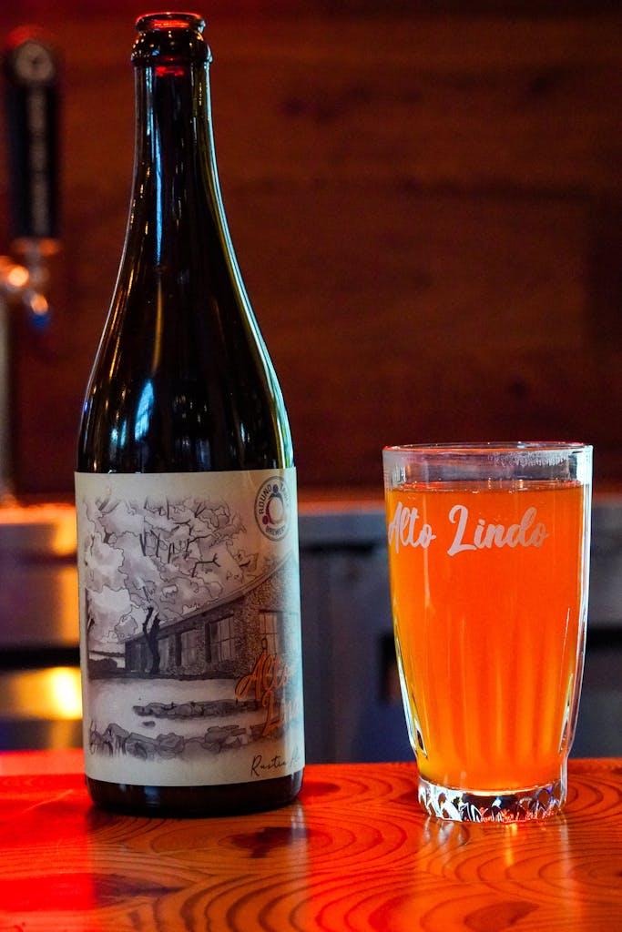 A tall bottle of Alto Lindo beer next to a glass filled with Alto Lindo beer and Alto Lindo written on the top of the glass. Red from the neon sign in the background backlighting the beer.