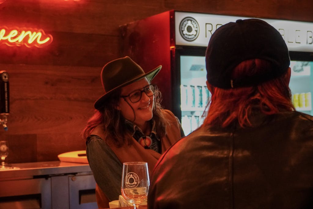 A person behind the bar with a western hat, smiling, leaning on the bar table talking their friends.