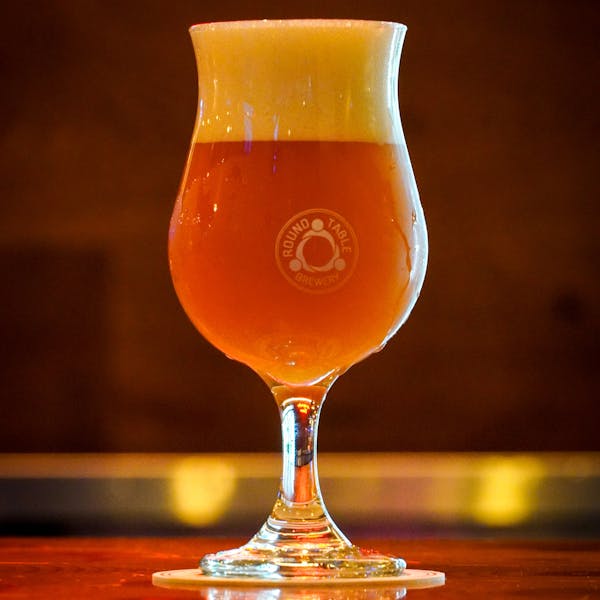 A hazy orange beer with foam in a Round Table Brewery glass.