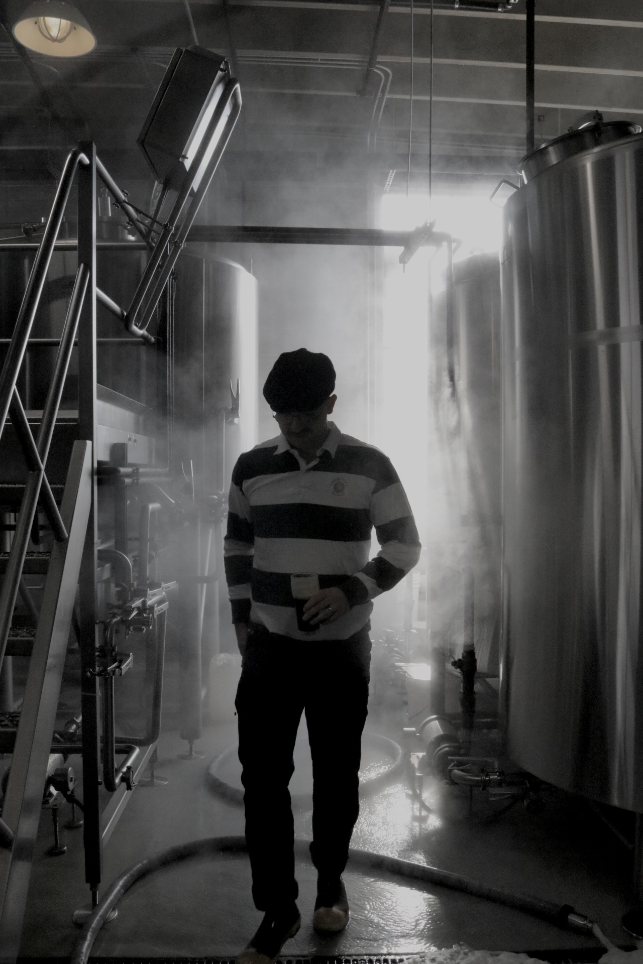 Picture of a man holding a beer in the distance with a brewery scene in the background with steam