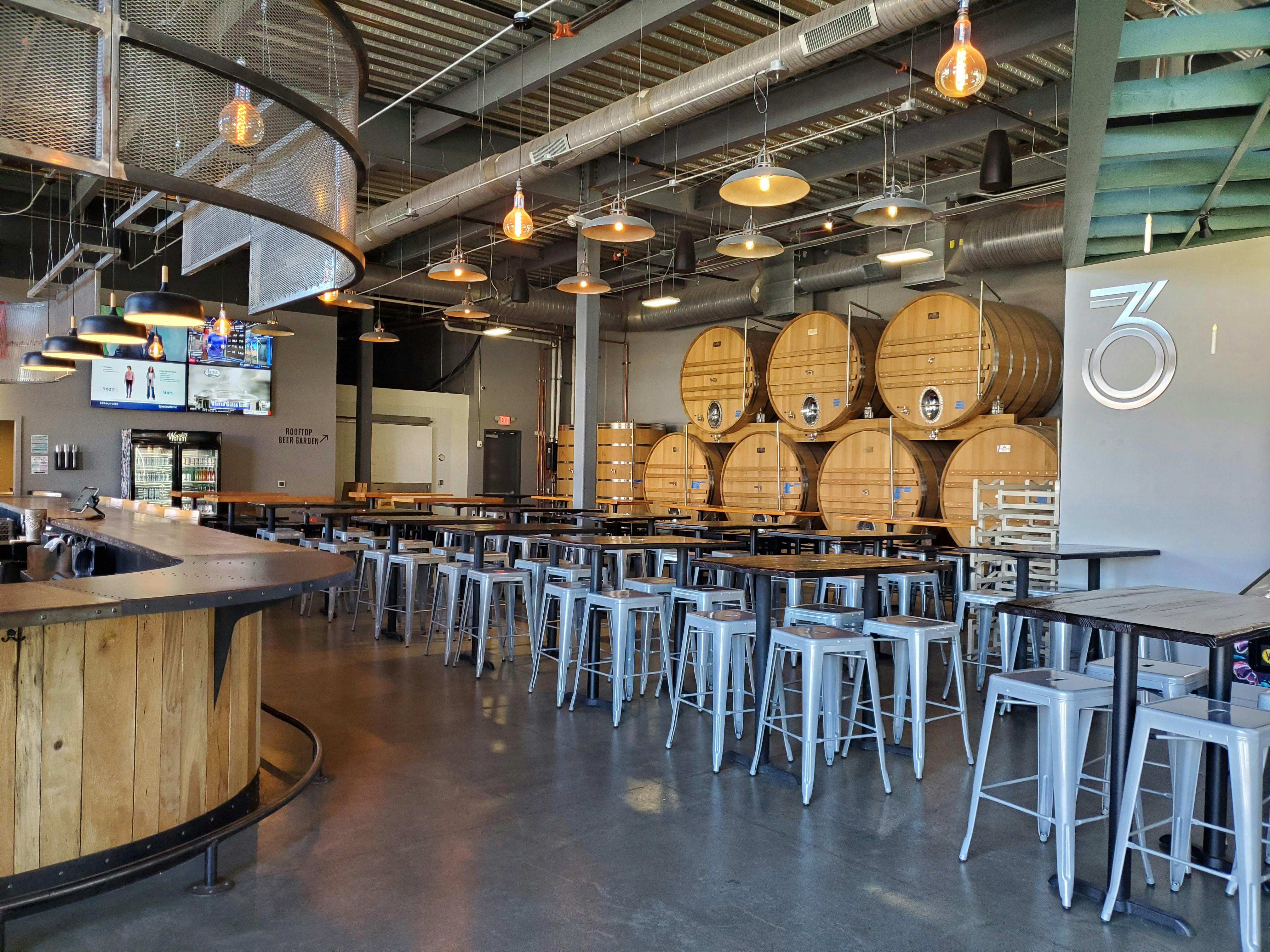 Taproom filled with 4-top tables, TVs on the wall in the background, and foeders along the back wall