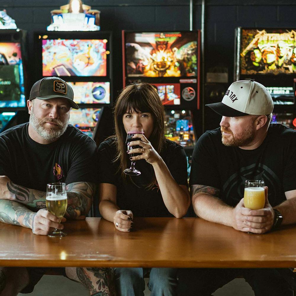 Brad, Seth, and Tara sitting at a table with beers in front of them and pinball machines in the back.